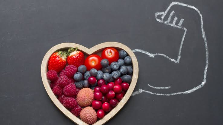 A heart-shaped bowl filled with fresh fruits like strawberries, blueberries, raspberries, lychees, cranberries, and tomatoes, placed on a black background with a chalk drawing of a flexing arm muscle, symbolizing strength and health through nutrition.