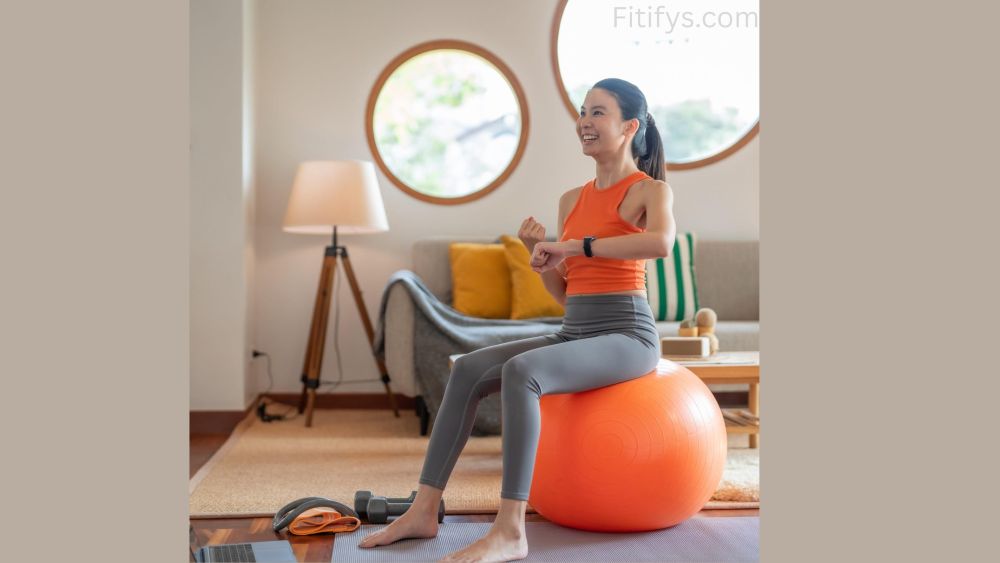 Smiling woman in workout clothes sitting on an orange stability ball at home, checking her smartwatch. She is in a cozy living room with a round window, couch, and lamp in the background. Dumbbells, resistance bands, and a laptop are nearby, indicating a home workout setup.