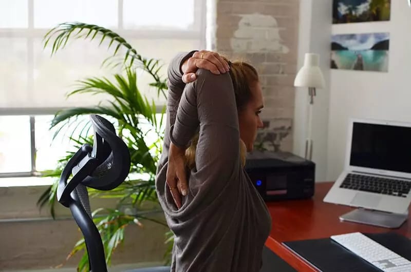 A professional woman in a cozy, modern home office is stretching at her desk. She sits in an ergonomic chair, arms raised behind her head in a tricep stretch. Natural light pours in through a large window with blinds, casting a warm glow on the room. A leafy green plant sits beside the desk, and the workspace features a laptop, keyboard, notebook, and a desk lamp. Wall decor includes travel or nature photos. The setting feels calm, productive, and wellness-focused. Style: realistic or high-quality lifestyle photography.