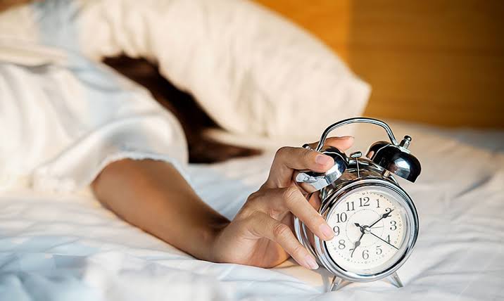 A person lying in bed reaches out with one hand to turn off a classic silver analog alarm clock, suggesting an early morning wake-up.
