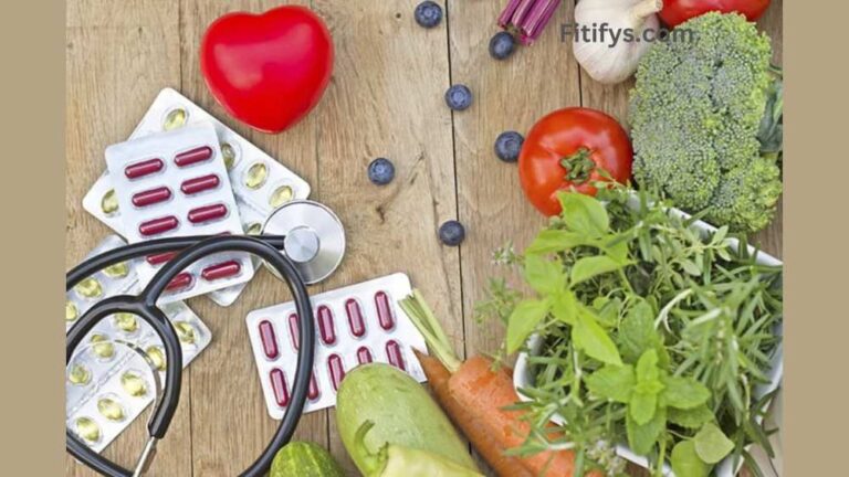 A flat lay image of a wooden table displaying heart health-related items: packs of red and yellow capsules, a stethoscope, a red heart-shaped object, fresh vegetables (broccoli, tomato, carrot, zucchini), leafy herbs, garlic, and scattered blueberries. The top right corner shows the branding "Fitifys.com."