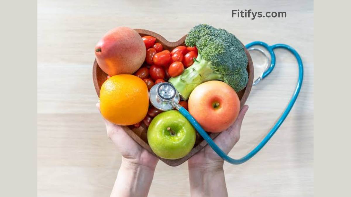 A pair of hands holding a wooden heart-shaped bowl filled with healthy fruits and vegetables, including an orange, green apple, red apple, peach, cherry tomatoes, and broccoli. A stethoscope is placed across the bowl, symbolizing heart health. The background is light wood, and “Fitifys.com” is visible at the top.