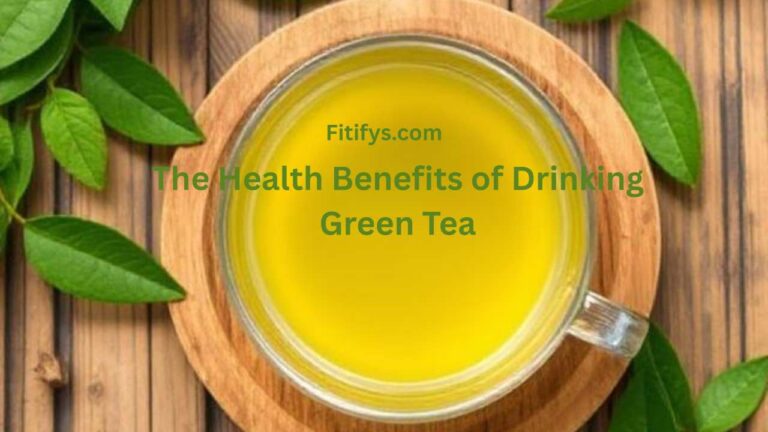 A clear glass cup filled with bright yellow-green tea placed on a round wooden coaster, surrounded by fresh green tea leaves on a wooden table. Overlaid text reads “Fitifys.com – The Health Benefits of Drinking Green Tea” in green font.