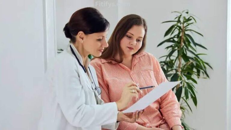 A female doctor in a white coat showing and explaining a document to a woman in a pink striped shirt, who listens attentively while sitting in a bright room with a potted plant in the background. The text “Fitifys.com” is faintly visible at the top.