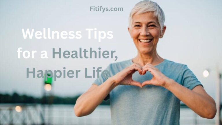 A smiling older woman with short gray hair stands outdoors, wearing a light blue T-shirt. She forms a heart shape with her hands in front of her chest. The background is softly blurred, showing a railing and sky. Overlaid text reads, “Wellness Tips for a Healthier, Happier Life,” with “Fitifys.com” above in smaller font.