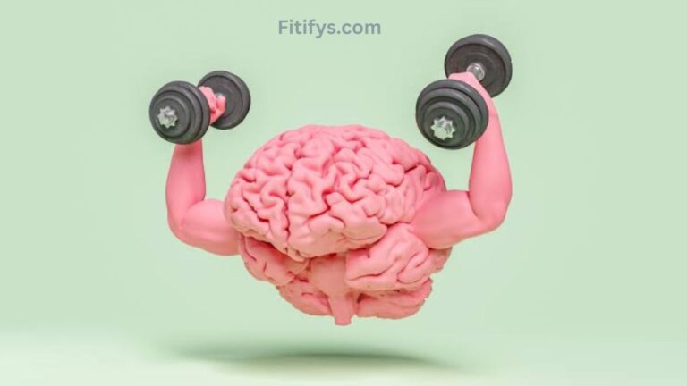 A 3D illustration of a human brain with muscular arms, holding dumbbells in each hand as if performing a bicep curl. The brain and arms are pink, while the dumbbells are black with silver accents. The background is a light pastel green, and the text “Fitifys.com” appears at the top.