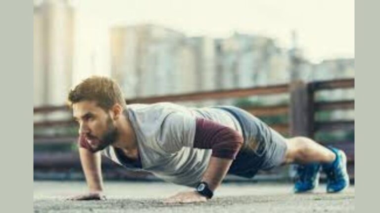A fit young man in athletic clothing performing a push-up outdoors on a concrete surface, with a focused expression. The background shows blurred urban buildings, giving the scene a city workout vibe.
