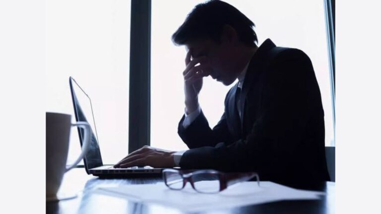 Stressed businessman sitting at his desk with a laptop, holding his head in frustration, symbolizing workplace stress and mental health challenges.