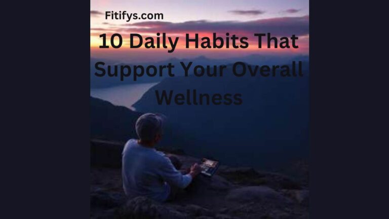 Person sitting on a rocky mountain ledge at sunset, holding a tablet and looking out over a scenic view of mountains and water. Text over the image reads “Fitifys.com – 10 Daily Habits That Support Your Overall Wellness” in black font.