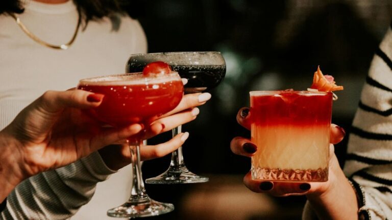 Three people raising colorful cocktails in a celebratory toast, featuring red, black, and layered orange-red drinks in elegant glasses.