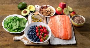 Healthy food assortment including fresh salmon, spinach, avocado, apples, blueberries, strawberries, nuts, flax seeds, and olive oil displayed on a wooden table, representing a balanced diet for wellness.